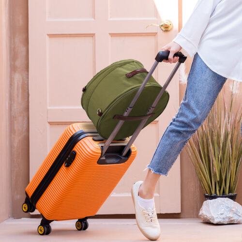 Young solo traveling woman standing with Georgian door as she is towing her bright orange suitcase filled with remote working tech, putting away plane tickets in her left pocket